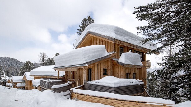 Eine Holzhütte mit schneebedecktem Dach und mehreren Balkonen, umgeben von schneebedeckten Bäumen und einer Bergkulisse.