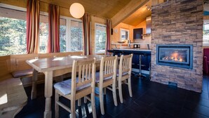 Ein gemütliches Blockhaus-Interieur mit einem Holzesstisch, vier Stühlen, einem Kamin aus Stein und einer Küchenzeile mit Herd und Schränken.
