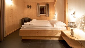 Quarto de madeira com cama, uma cadeira, janela com cortinas e abajur em uma mesinha de cabeceira.
