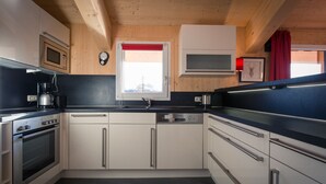 A modern kitchen with wooden ceiling beams, a window with a red blind, stainless steel appliances, and a black countertop. - Chalet #40 with IR-sauna and indoor whirlpool (Turrach)