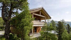 A two-story wooden cabin with a balcony, surrounded by trees and mountains in the background. - Chalet #40 with IR-sauna and indoor whirlpool (Turrach)