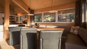 Een modern interieur met een houten plafond, een eethoek met stoelen en een tafel, een bank en grote ramen met uitzicht op bomen.