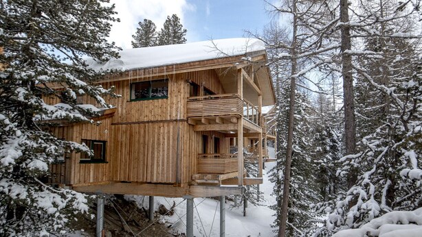 Une cabane en bois, surélevée sur des piliers, se dresse au cœur d’une forêt enneigée.