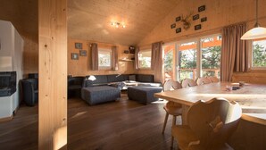 Un intérieur de chalet chaleureux avec un plafond en bois, un coin repas doté d’une grande table et de chaises, un coin salon avec un canapé et un pouf, et une cheminée.