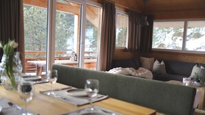 A dining area with a wooden table set for a meal, a green sofa in the background, and large windows offering a view of trees.