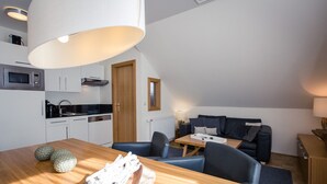 A modern kitchen with white cabinets, a microwave, and a stove. There is a wooden dining table with a bowl and decorative items. A dark sofa and a lamp are in the living area. - Apartment # 12 with balcony (Turrach)