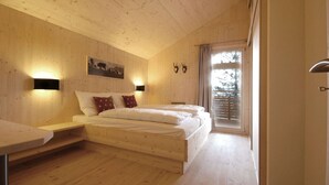 Une chambre avec un plafond en bois, un lit avec une literie blanche, une table de chevet, une lampe fixée au mur, une fenêtre avec des rideaux et un tableau accroché au mur.
