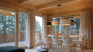 Un intérieur en bois avec une table et des chaises, de grandes fenêtres avec des rideaux et une vue sur les arbres à l’extérieur.