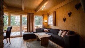 Ein gemütliches Wohnzimmer mit einem dunkelgrauen Sofa, einem Holztisch und einem Teppich mit Muster. Große Fenster mit Vorhängen lassen natürliches Licht herein und bieten Ausblick ins Freie.