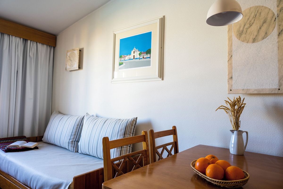 A neatly arranged room with a bed, a dining table with a basket of oranges, a vase with dried plants, and a framed picture on the wall.