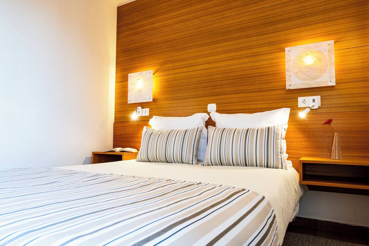 A hotel room with a wooden headboard, a large bed with striped bedding, two bedside tables with lamps, and a wall-mounted light fixture.
