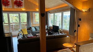 Un soggiorno accogliente con divano, tavolino da caffè e televisione. Sono presenti ampie finestre con tende e travi a vista sul soffitto.