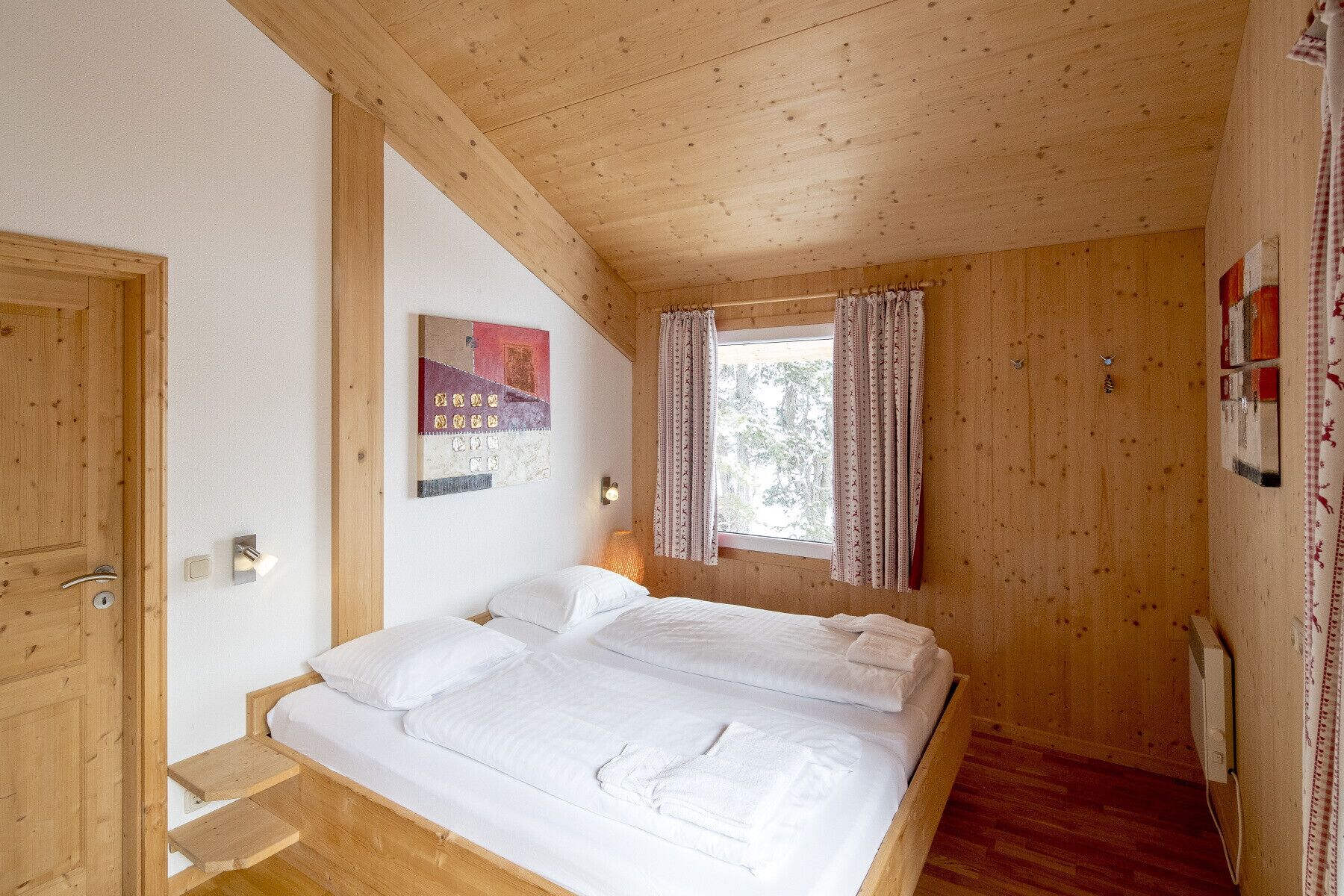 A wooden room with a bed, a window with curtains, a wall-mounted light, and a door.