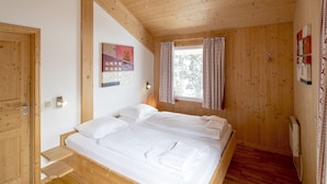 Una stanza in legno con un letto, una finestra con tende, una lampada a parete e una porta.