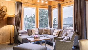 A cozy room with a sectional sofa, a small table, and a lamp. Large windows offer a view of trees and mountains.