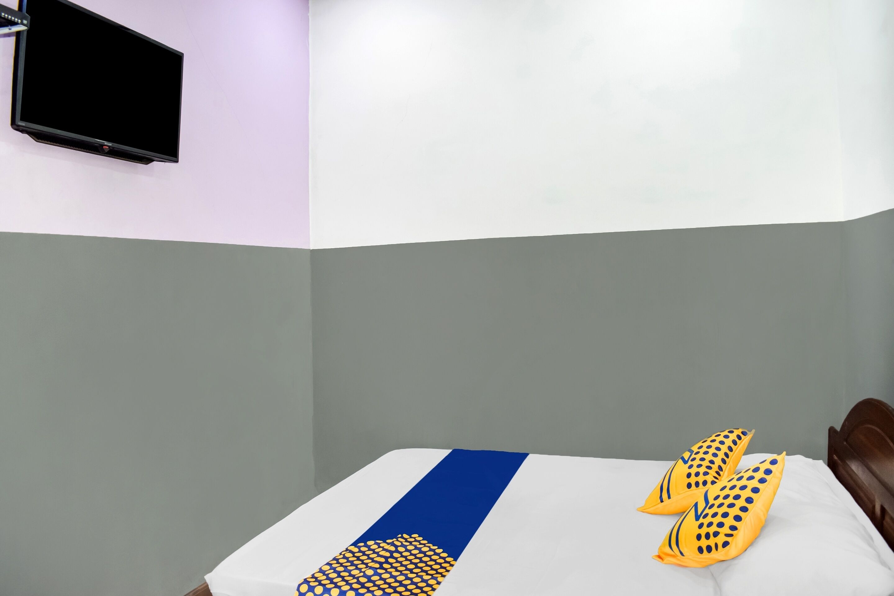 Habitación de hotel con una cama, un televisor de pantalla plana montado en la pared y una almohada decorativa azul y amarilla sobre la cama.