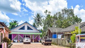 A rural setting with a house featuring a blue roof and a purple facade, a white car parked in front, and a small truck. - Hotel O Penginapan Widodo (Sebulu)