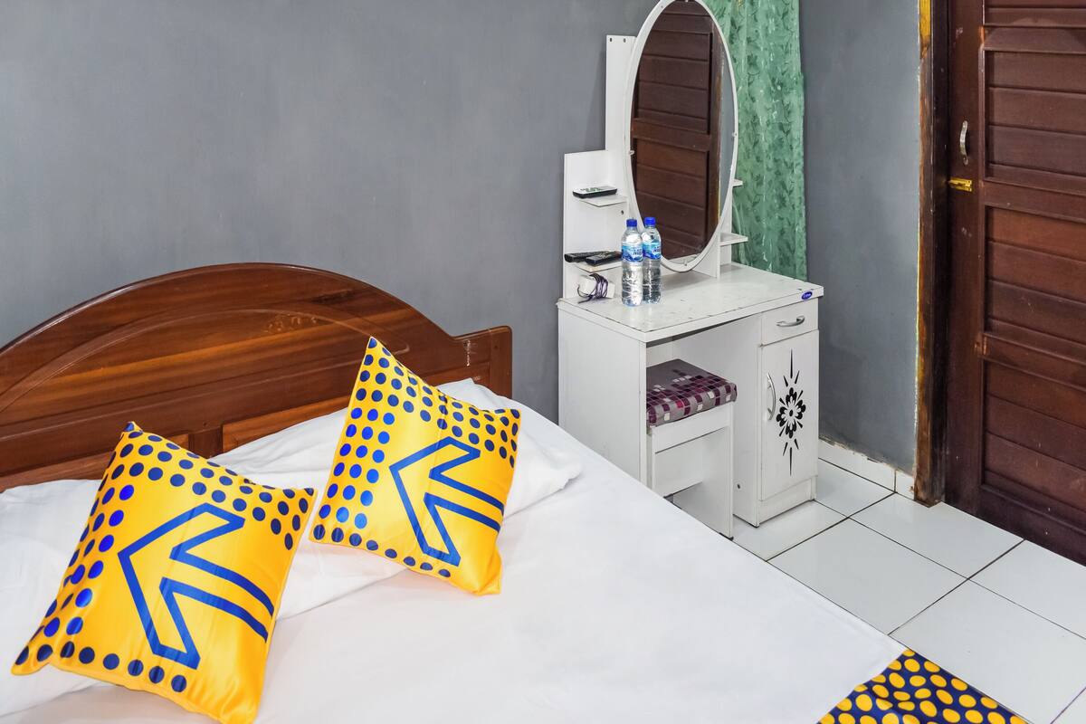 A bedroom with a wooden bed, two pillows with geometric patterns, a white bedside table with a mirror, a water bottle, and a green curtain.