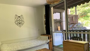 Una habitación compacta con un sofá blanco, una pequeña mesa de madera y un armario de madera. Se observa una obra de arte geométrica en la pared, y la habitación tiene vistas a un área exterior con terraza de madera y vegetación a través de una puerta de cristal.