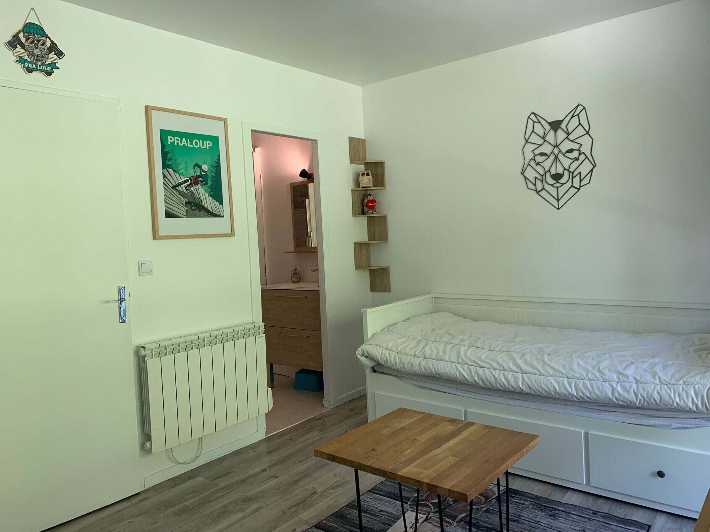 Une chambre à coucher avec un lit, une table en bois, un radiateur, une affiche encadrée et un motif géométrique sur le mur.