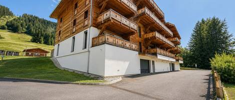 Un edificio de madera de varios pisos con balcones, un camino pavimentado y un área verde con árboles al fondo.