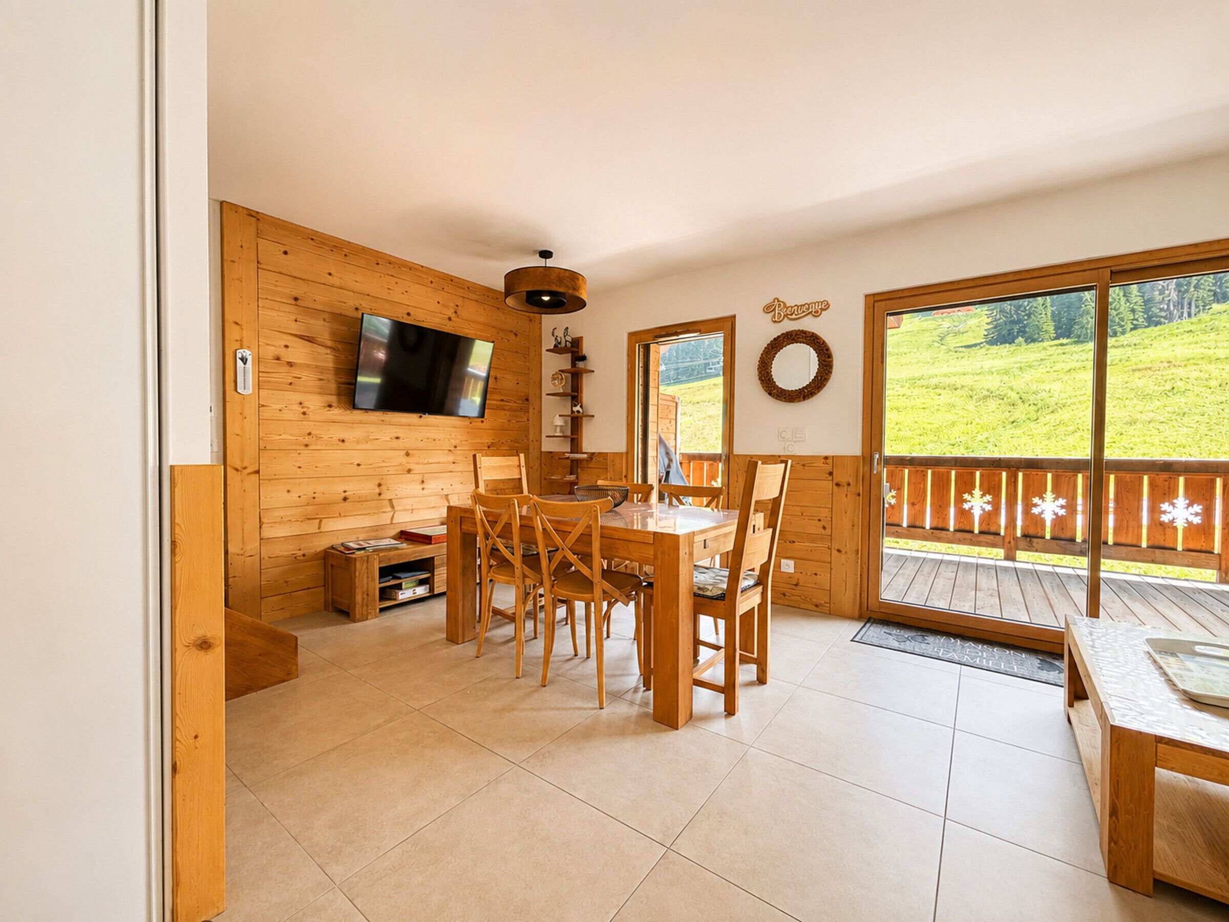 A wooden interior with a dining table and chairs, a flat-screen TV mounted on the wall, and a view of a green landscape through a glass door.