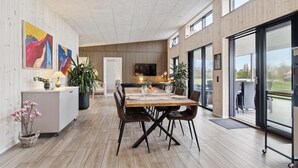 A modern dining area with a wooden table and chairs, a TV mounted on the wall, and large windows offering a view of the outdoors. - Gorgeous home in Hadsund with sauna (Hadsund)