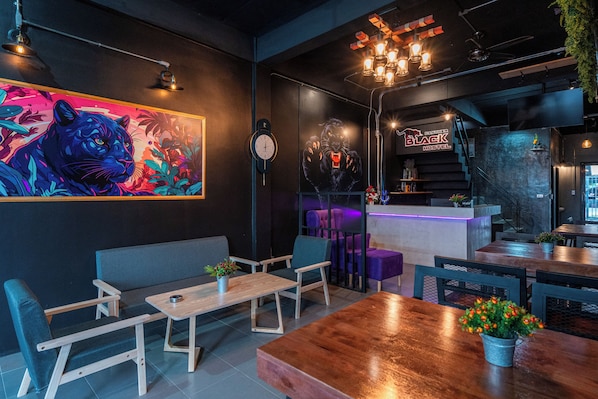 A modern interior with a large mural, wooden furniture, and a staircase leading to a bar area. - Black Panther Hostel (Phuket)