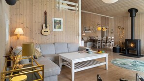 A cozy living room with a wooden interior, a sofa, a coffee table, a fireplace, and a dining area with chairs and a table. - Amazing home in Fårevejle with sauna (Fårevejle)
