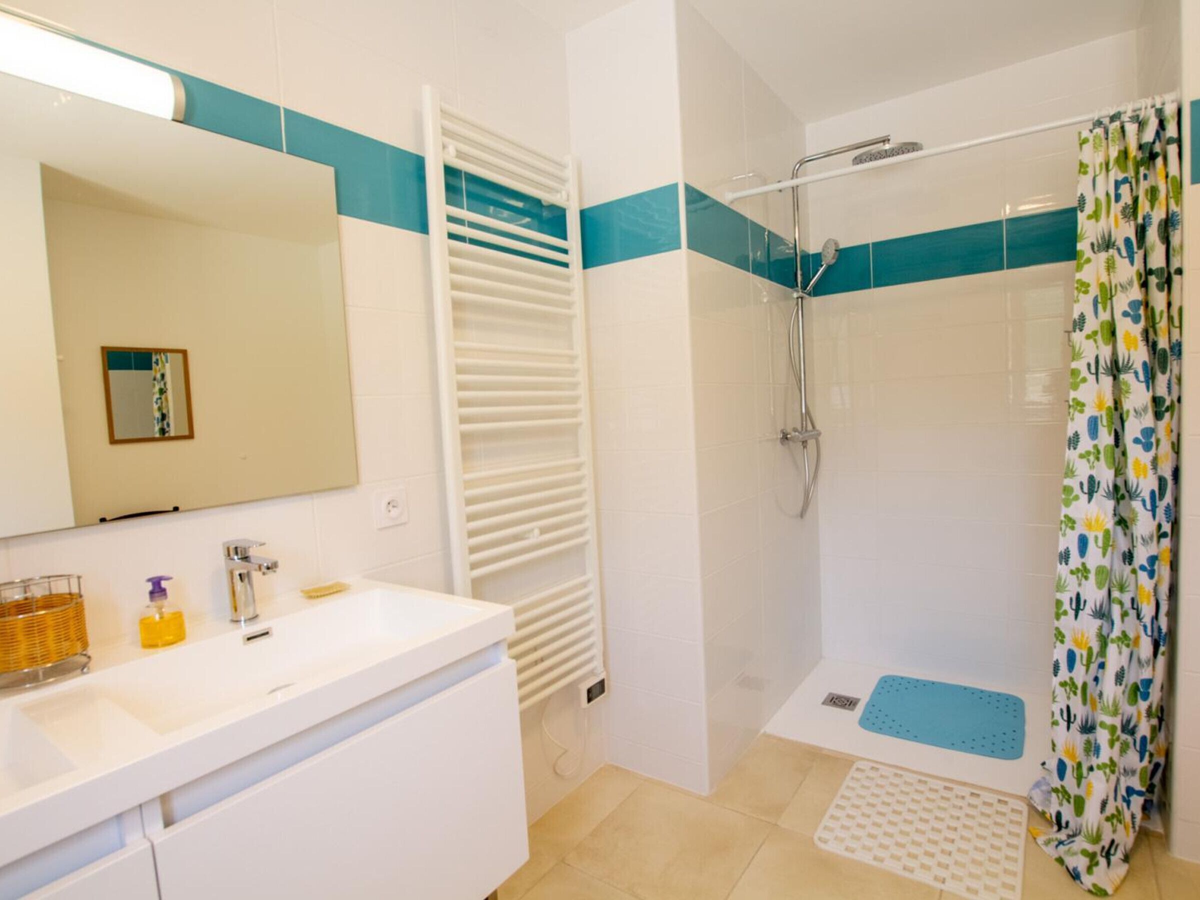 Une salle de bain avec un lavabo blanc, un miroir et un porte-serviettes. On y trouve une douche avec une porte en verre et un rideau de douche orné d’un motif de cactus.