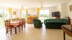 Een lichte, ruim opgetogen kamer met een eettafel en stoelen, een groene bank, een televisie en een keukenhoek.