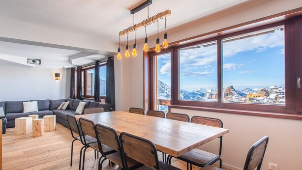 Un espace repas moderne avec une table et des chaises en bois, un canapé et une grande fenêtre donnant sur les montagnes.