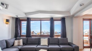Un salon moderne avec un canapé d’angle, des tables d’appoint en bois et de grandes fenêtres offrant une vue sur les montagnes.