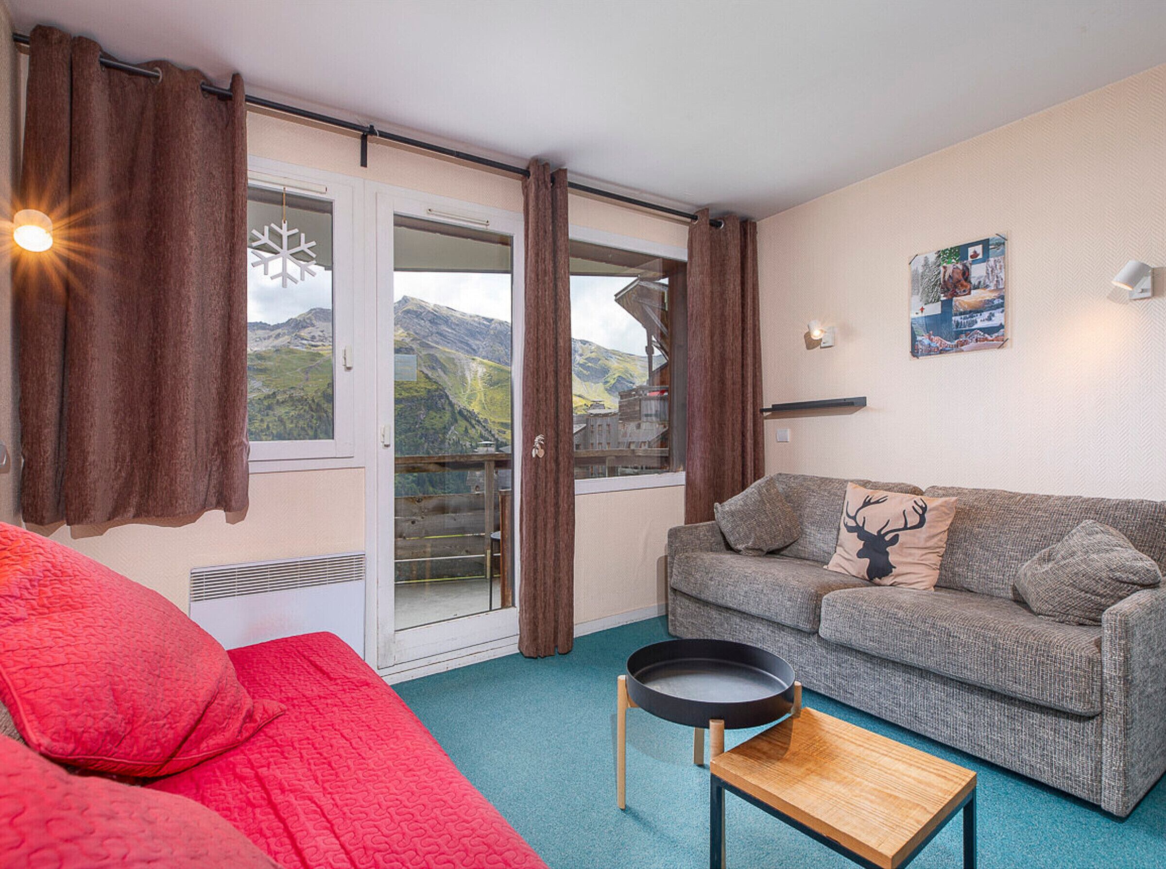 Ein gemütliches Wohnzimmer mit einem grauen Sofa, roten Kissen, einem Holztisch und Bergblick durch eine Glastür.
