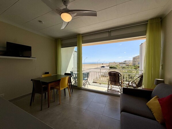 Ein Zimmer mit Esstisch, Stühlen, einer Couch, einem Fernseher und einem Balkon mit Blick auf den Strand.
