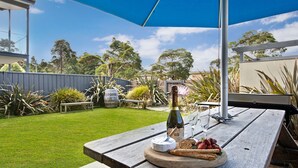 A picnic setup with a bottle of champagne, glasses, a cheese wheel, crackers, and strawberries on a wooden table under a blue umbrella. - Serenity Shores by Book Jervis Bay (Huskisson)