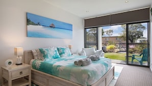 A bedroom with a large bed, a nightstand, a painting of a beach, and a view of the outdoors through a sliding door. - Interlude Apartment by Book Jervis Bay (Huskisson)