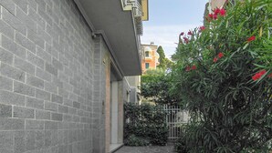 Une ruelle étroite, avec un mur de briques d’un côté et un immeuble doté d’un balcon de l’autre. On y trouve une porte et quelques plantes vertes ornées de fleurs rouges.