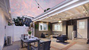 Un espace extérieur aménagé en patio, avec une table à manger, des chaises et un canapé. Des guirlandes lumineuses sont suspendues au-dessus, et une clôture blanche se trouve au fond.