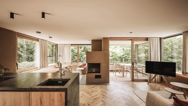 Une cuisine moderne dotée d’un grand îlot central, d’une cheminée et d’un coin repas offrant une vue sur la forêt.
