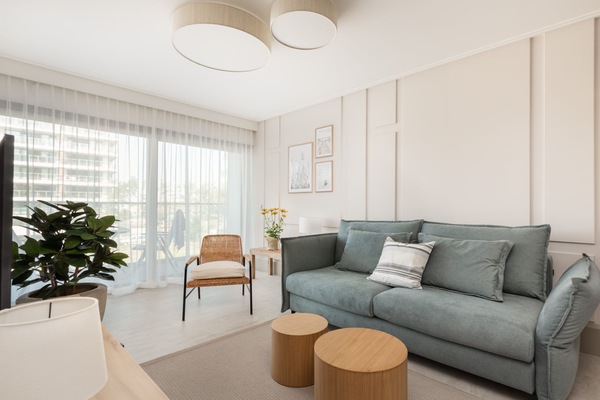 A bright living room with a modern design, featuring a comfortable gray sofa and large windows overlooking greenery.