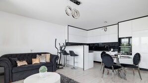 Una sala de estar moderna con un sofá negro, una mesa de centro blanca y una pequeña planta en maceta. Hay una zona de comedor con una mesa y sillas, y una cocina con armarios blancos y electrodomésticos negros.