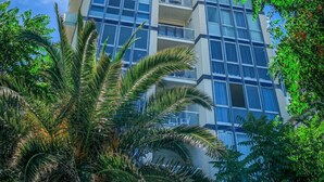 A modern multi-story building with large windows and balconies, surrounded by lush greenery and palm trees. - Luxury Suites Collection-Viale Milano 3 (Riccione)