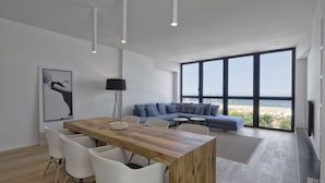 A modern living room with a wooden dining table, white chairs, a blue sofa, and large windows offering a view of the city. - Luxury Suites Collection-Viale Milano 3 (Riccione)