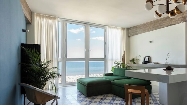 A modern living room with a green sectional sofa, a small wooden chair, a white kitchen island, and a large window offering a view of the beach. - Luxury Suites Collection-Viale Milano 3 (Riccione)