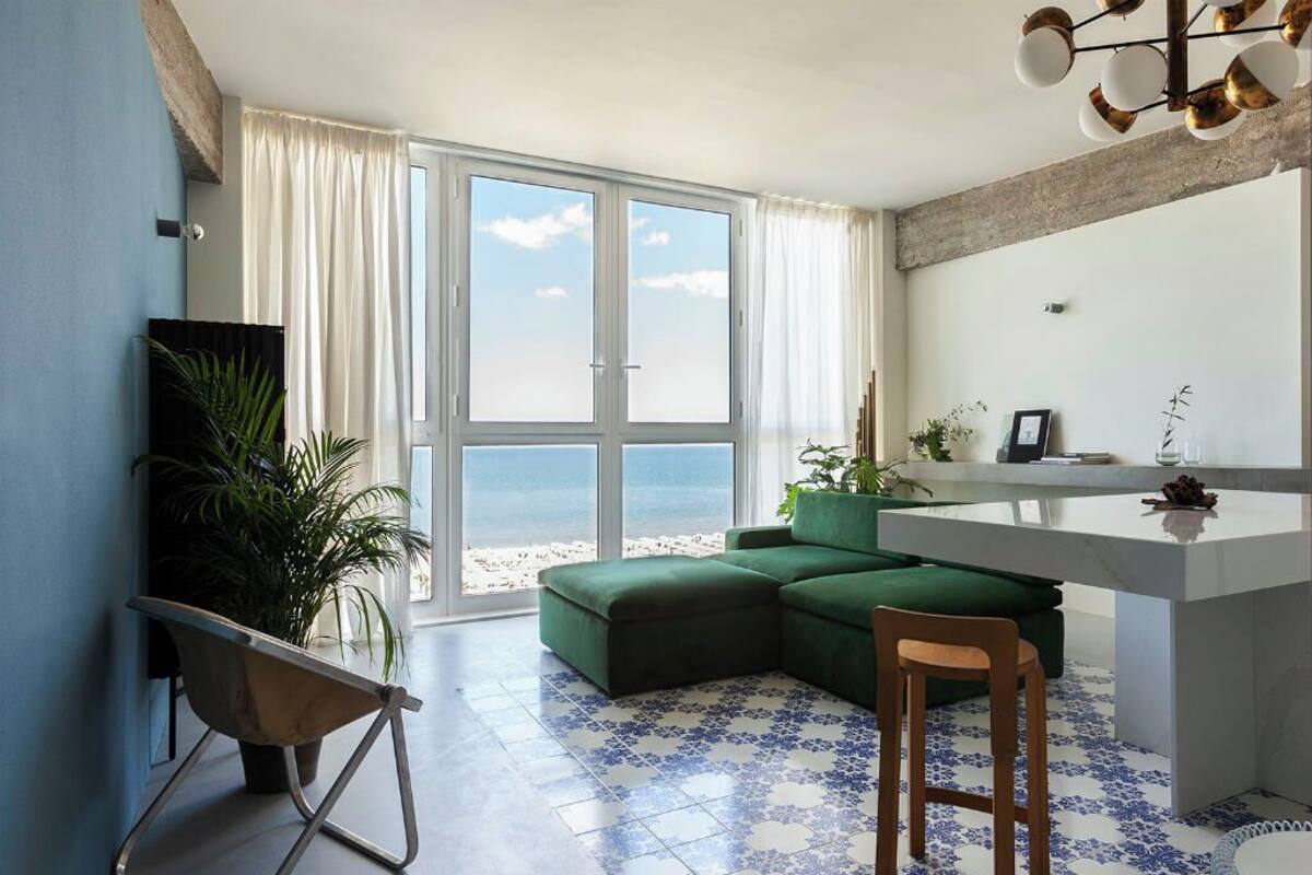 A modern living room with a green sectional sofa, a small wooden chair, a white kitchen island, and a large window offering a view of the beach.