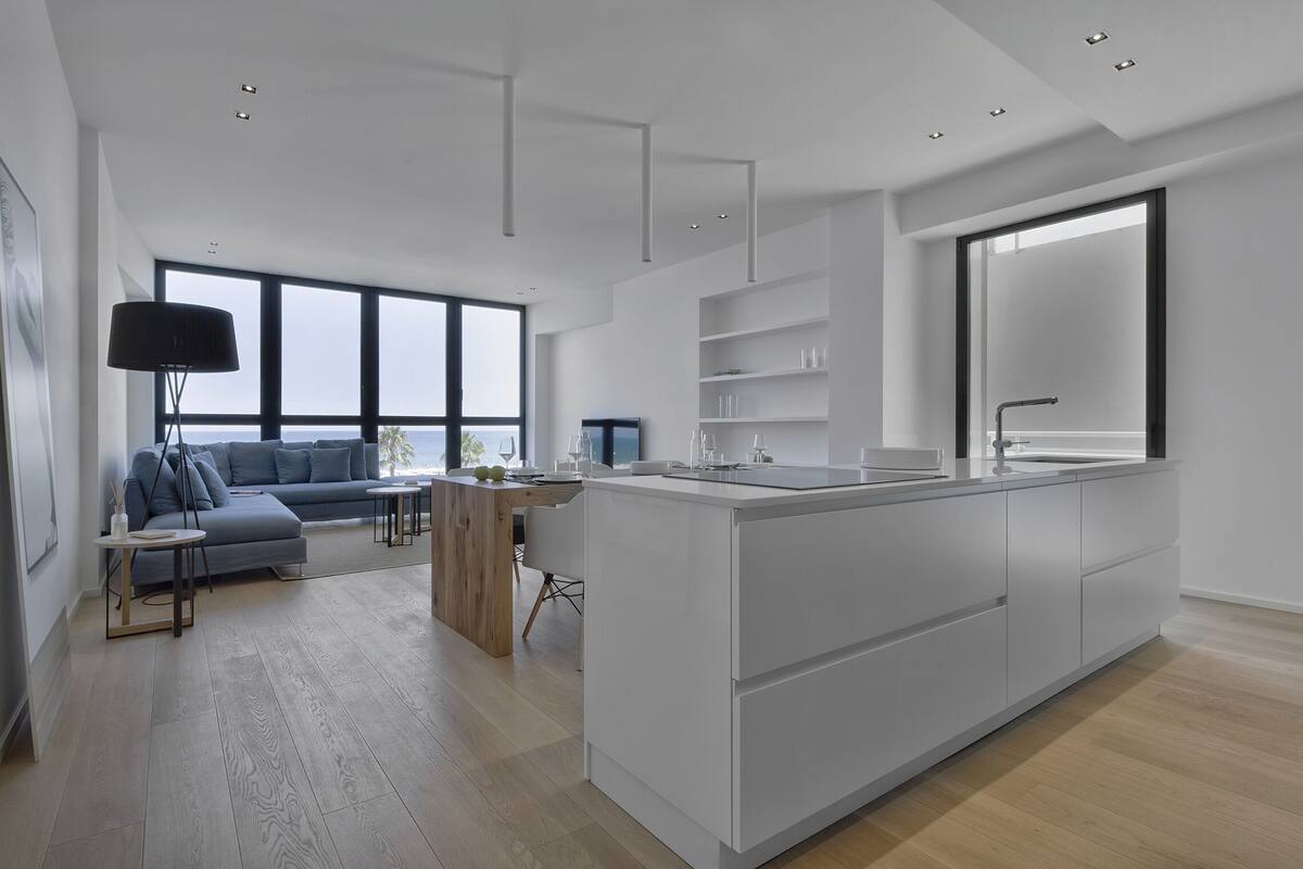 A modern kitchen with a central island, a dining area with a sofa and table, and a large window overlooking the sea.