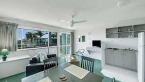 A modern kitchen with white cabinets, a built-in oven, and a refrigerator. There is a dining area with a table, chairs, and a window with curtains. - Reef Adventureland Motel (Tannum Sands)