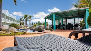 A poolside area with lounge chairs, a covered seating area, and a building with a white facade and green roof. - Reef Adventureland Motel (Tannum Sands)