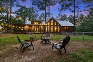A rustic cabin with a fire pit and outdoor seating, surrounded by trees and string lights. - New Listing · Tucker`d Out · Foosball, Hot Tub, & Prime Location! (Broken Bow)